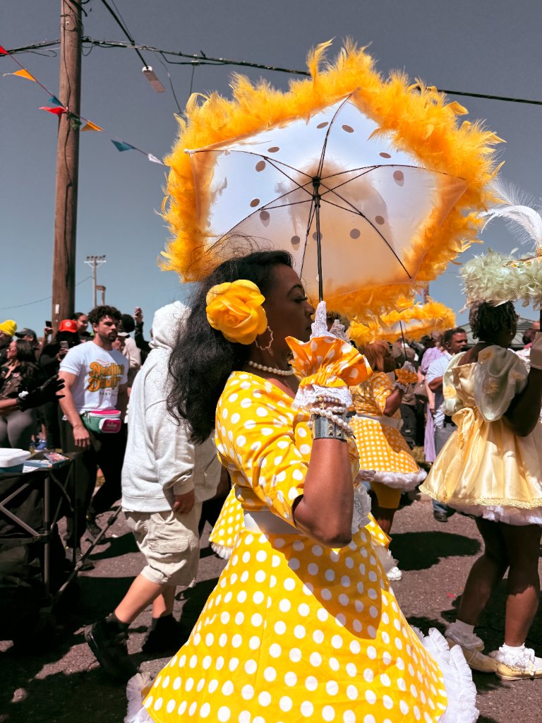 A woman in a vibrant yellow polka dot dress holds a large golden feathered parasol at a New Orleans street celebration
