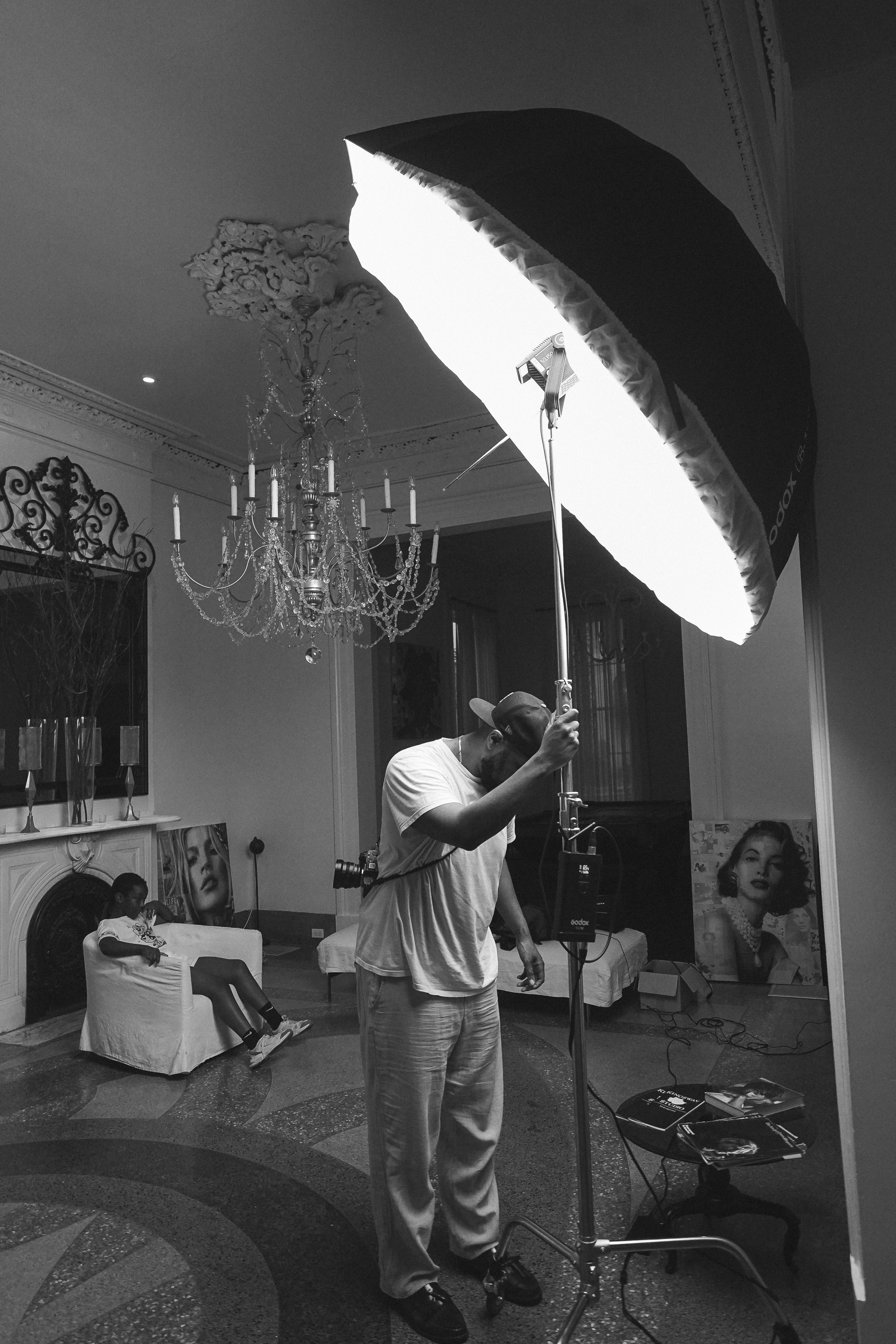 A behind-the-scenes shot of a photographer holding a studio softbox stand in an ornate room with a seated figure and framed art, black and white