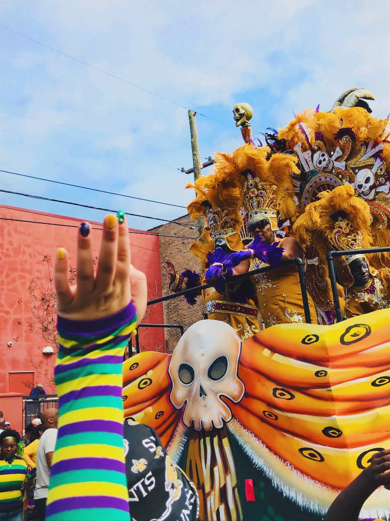 A colorful Mardi Gras float with vibrant feathers, a skull centerpiece, and a person with a rainbow-striped arm raised in celebration