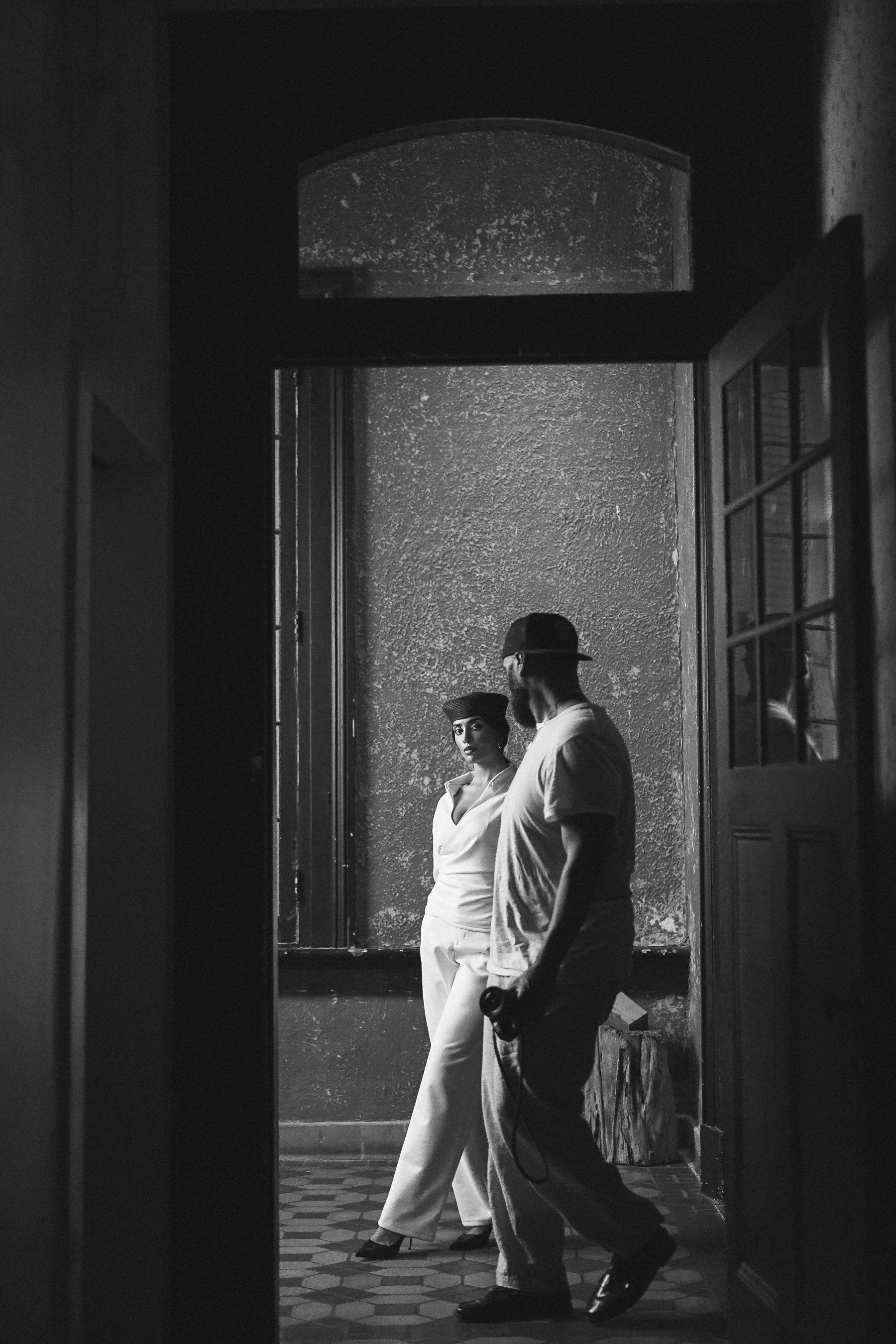 A woman in a white blazer and dark beret in a doorway looking back, with a photographer walking past in the foreground, black and white