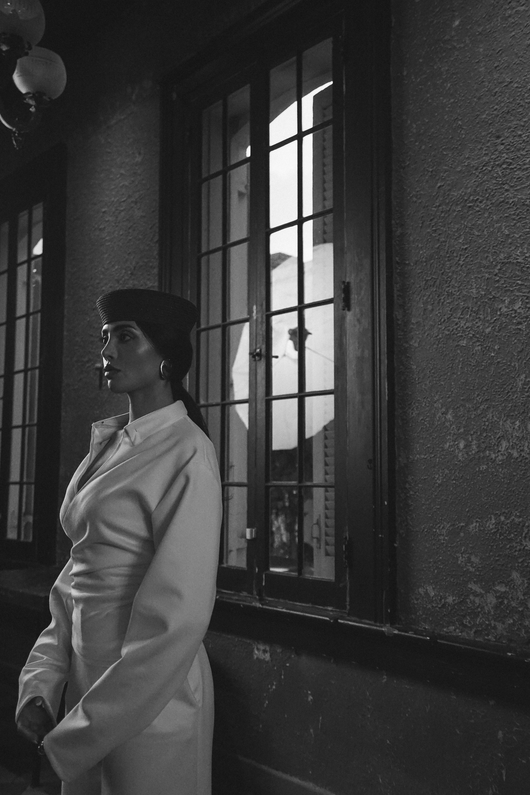 A woman in a dark pillbox hat and white collared shirt standing in profile near a dark-framed window, black and white portrait