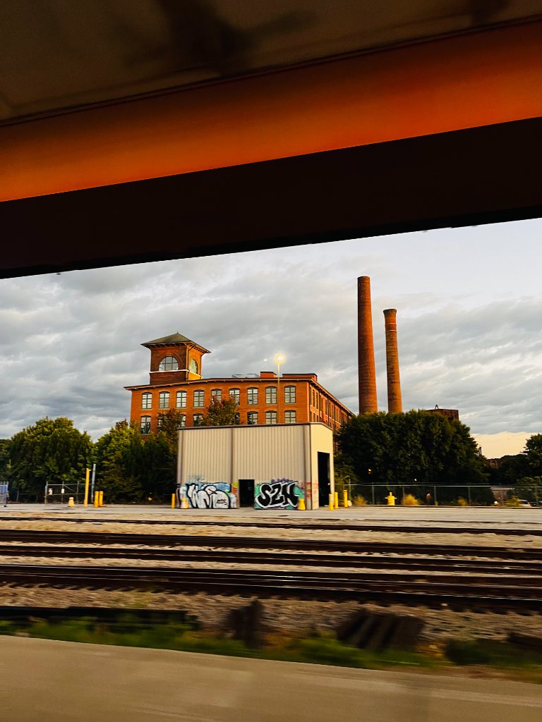 Railroad tracks leading past a graffiti-covered utility shed toward a historic brick industrial building with twin smokestacks, golden sunset light