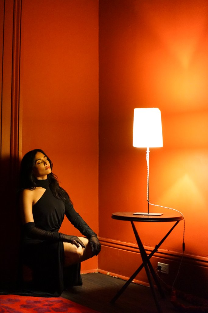 A woman in a black dress and gloves seated on the floor leaning against an orange wall, eyes closed, a lamp on a pedestal table casting warm light