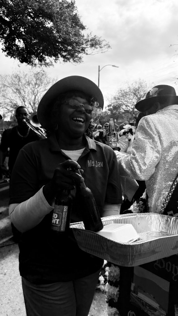 A smiling woman in a brimmed hat and glasses holding two beer bottles behind a catering tray at a street festival, black and white documentary style