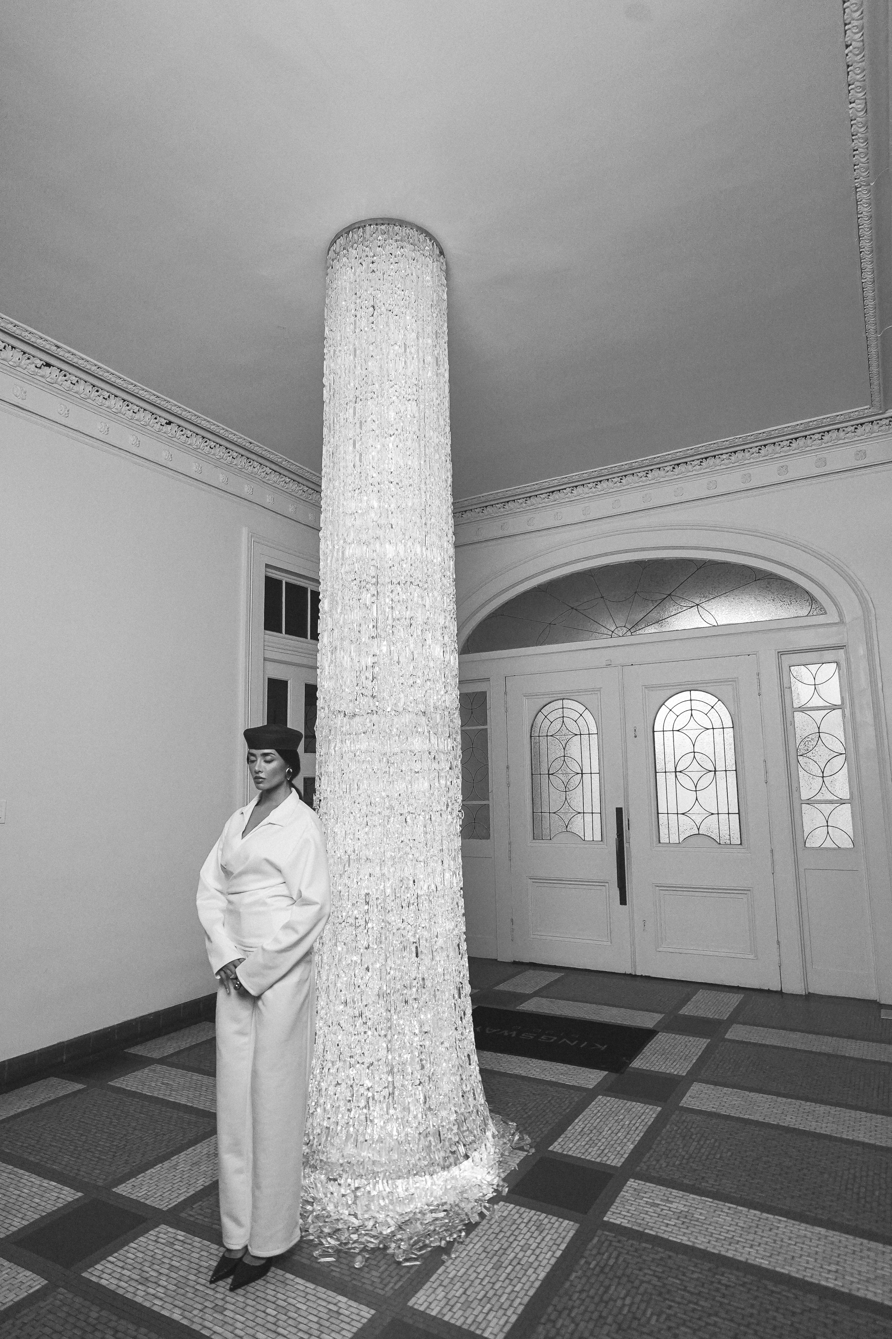 A person in a white wrap jacket and trousers with a dark beret standing beside a glowing cylindrical crystal chandelier column in a grand entrance hall, black and white portrait