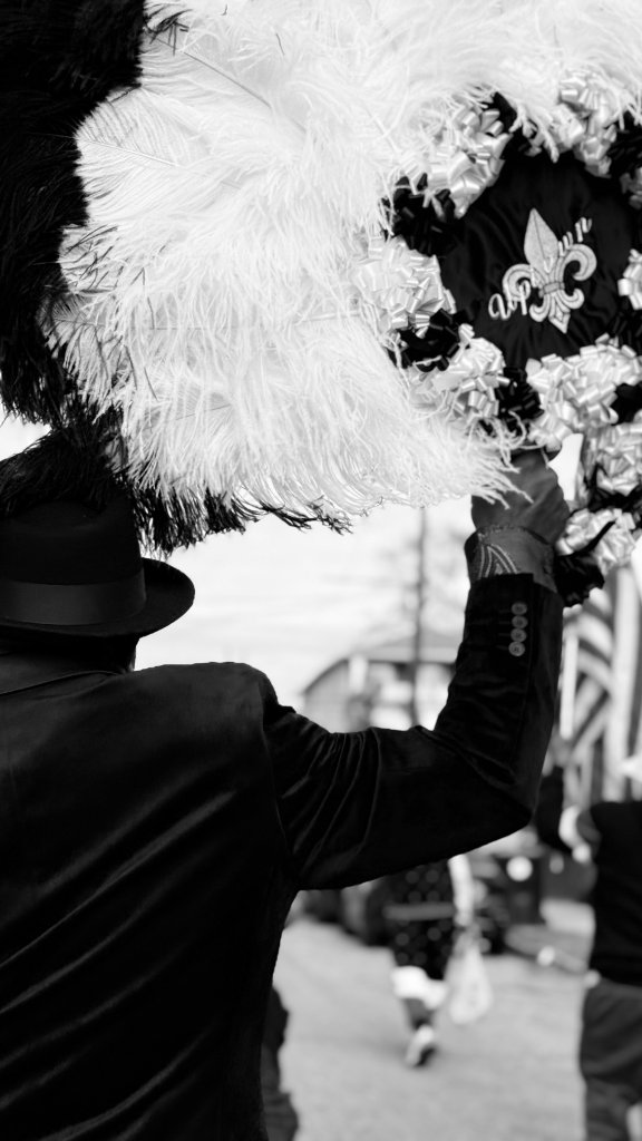 A parade participant in a dark fedora hat and jacket, arm raised high holding an ornate feathered emblem with a fleur-de-lis, black and white