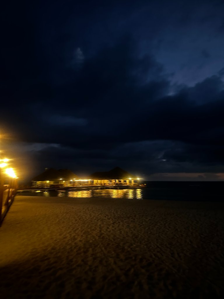 A nighttime beach pier lit by golden lights reflecting on calm water, dark dramatic sky above, sandy beach in foreground