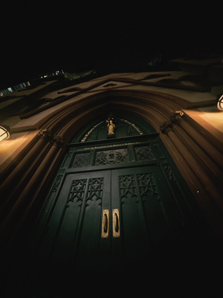 A dramatic upward view of an ornate green cathedral door with gold handles, lit by warm spotlights against dark night sky