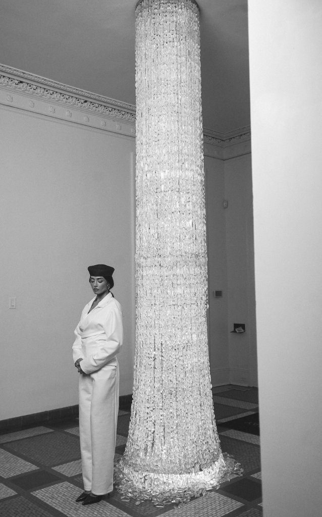 A person in a white long-sleeved garment and dark beret standing beside a tall crystal pillar in an interior setting with patterned tile floor, black and white