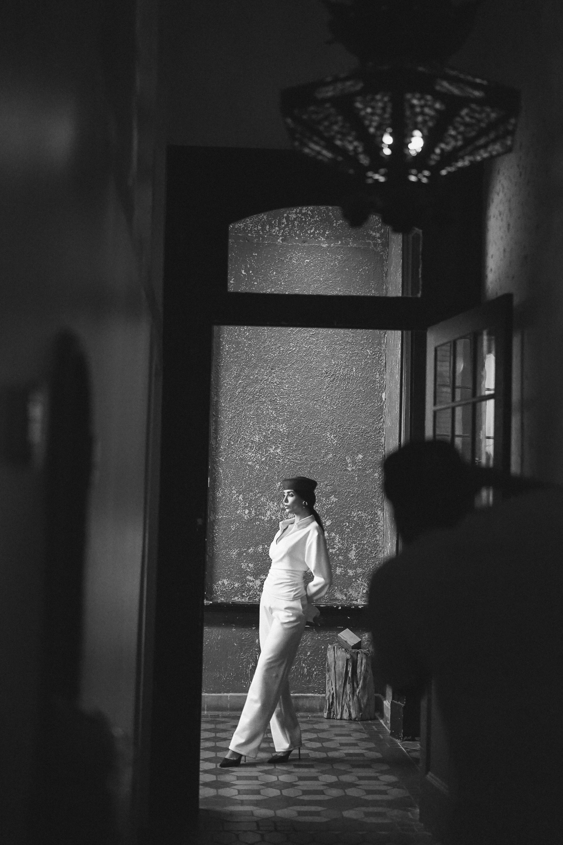 A model in a white outfit and dark hat standing in an arched corridor with a photographer silhouette in the foreground, black and white