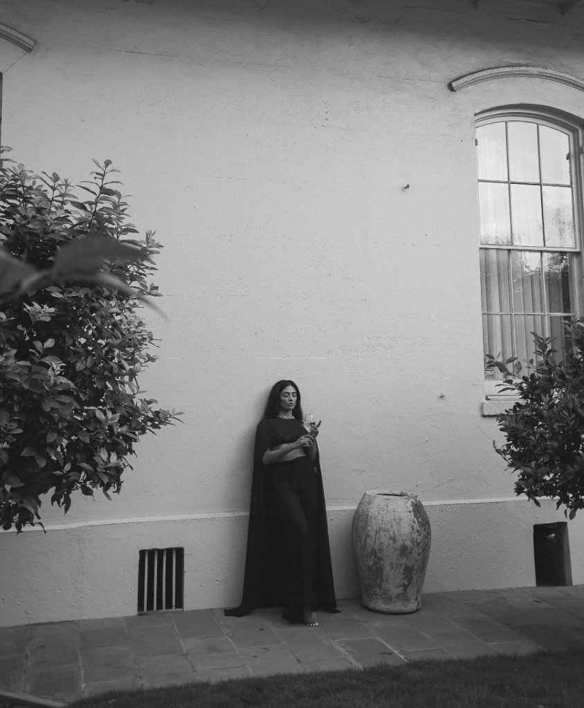 A woman in a dark ensemble with a cape holding a glass against an off-white wall, bushes and an arched multi-pane window, stone urn, black and white