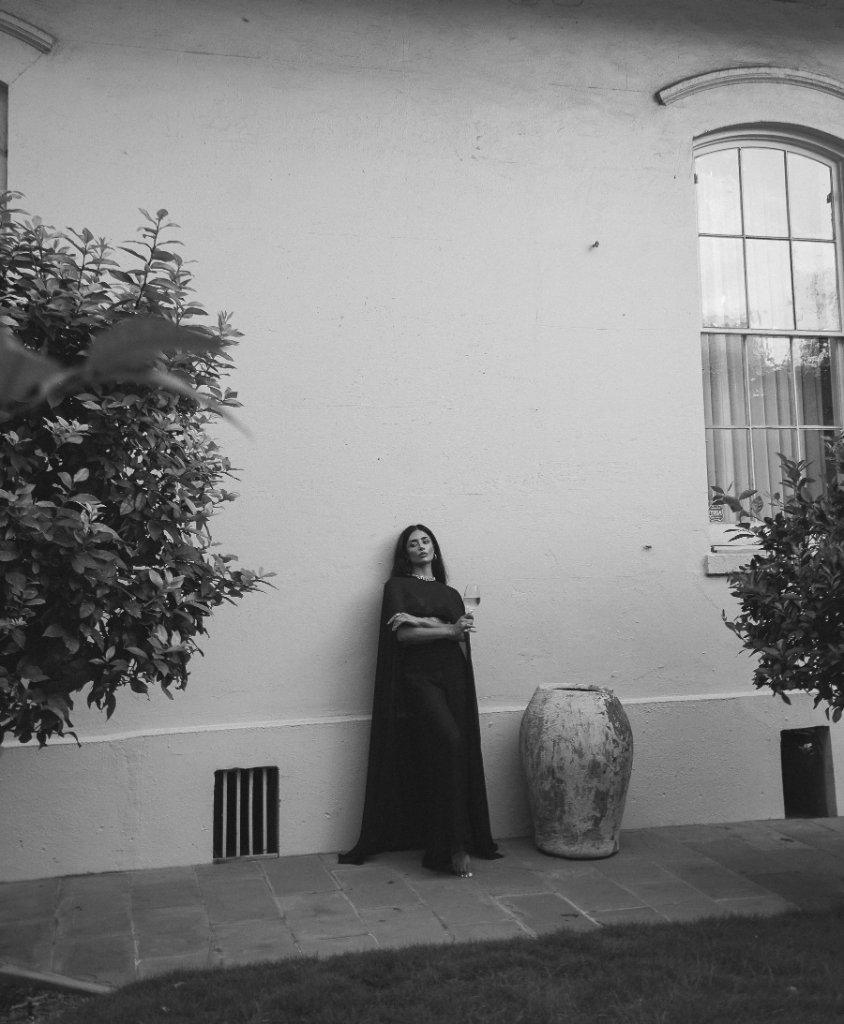 A woman in a dark long-sleeved outfit and flowing cape holding a wine glass against a light stucco wall with a multi-pane window, black and white portrait