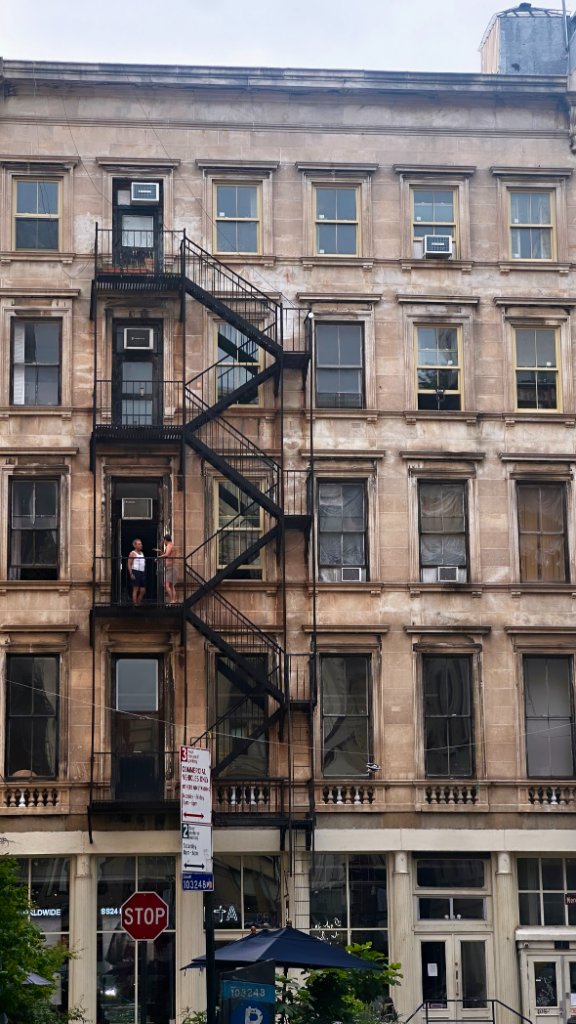 A tall urban building facade with a dramatic black metal fire escape zig-zagging down the sandstone wall, figures visible on the landing