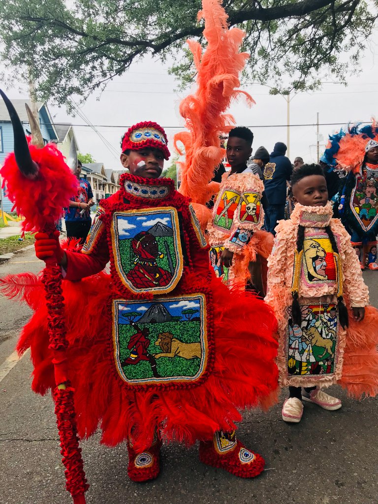 Three young children in elaborate Mardi Gras Indian costumes with feathered headdresses, beaded patches, standing on an asphalt street under cloudy sky