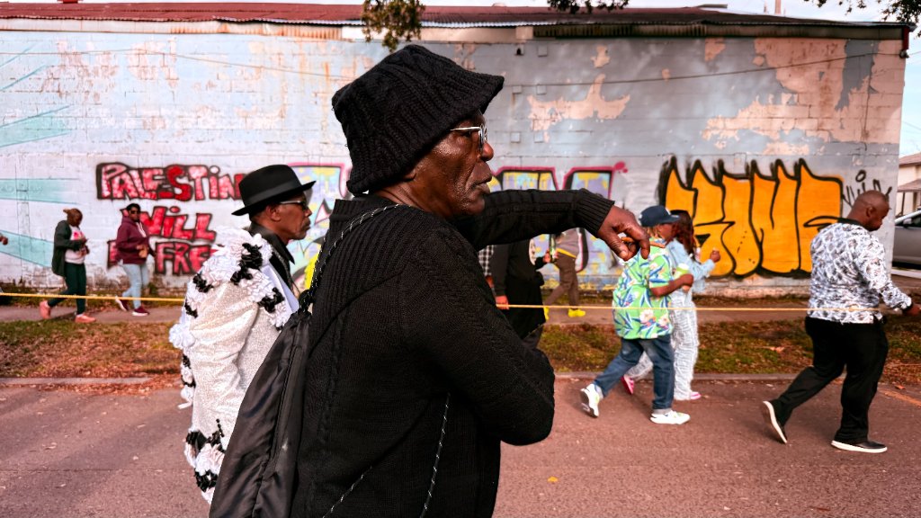 A man in a black beanie pointing ahead during a street parade, people walking past a graffiti-covered wall reading Palestine Will Free