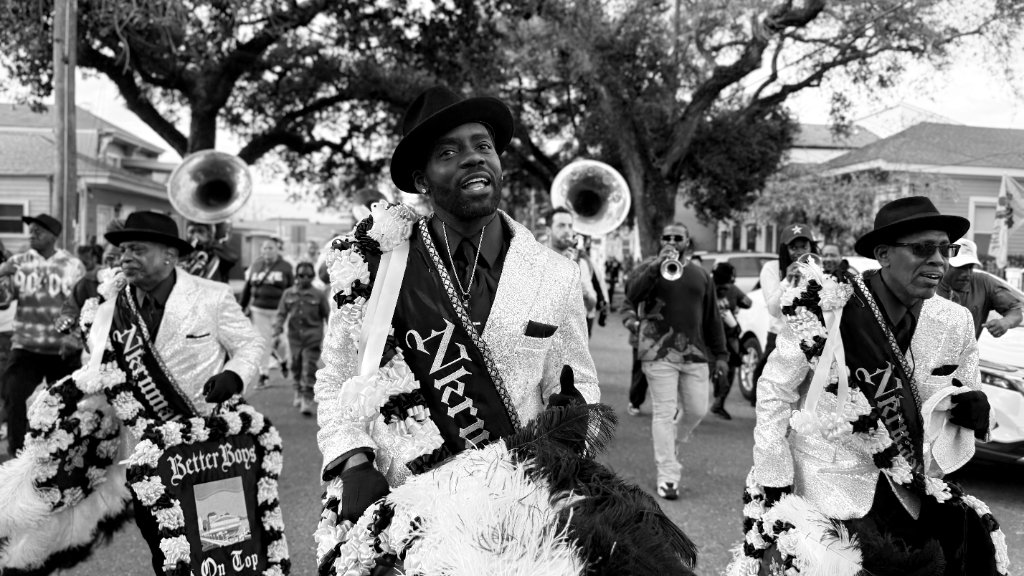 Three men in matching sequined jackets and dark fedora hats with Nkrumah sashes, marching in a street parade with a brass band visible behind them, black and white
