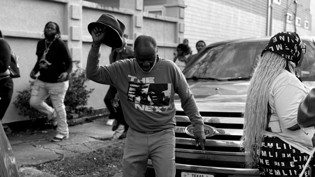 A street celebration with a man in the center lifting his hat, surrounded by people and a truck, black and white photography