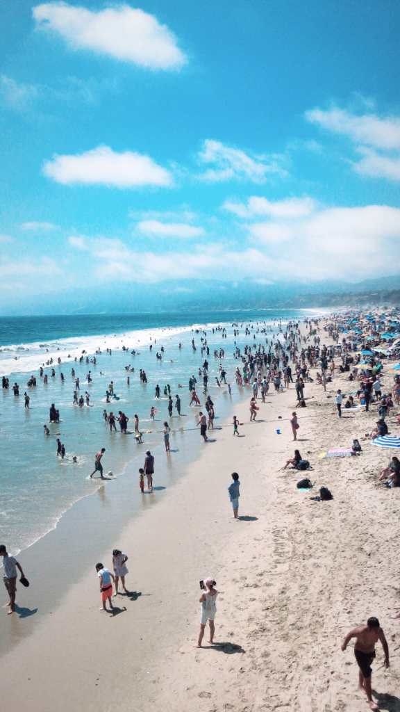A bright, sunny beach crowded with swimmers and sunbathers — turquoise water, white sand, blue sky with white clouds, wide overhead perspective