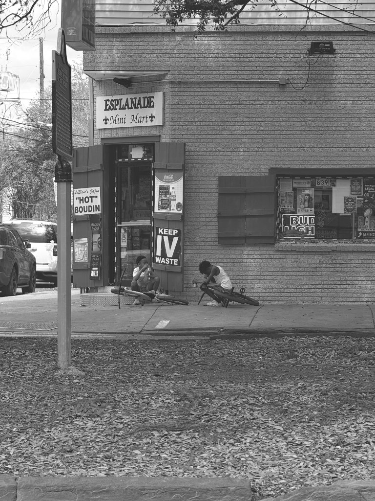 Two kids sit on the sidewalk outside the Esplanade Mini Mart with their bikes, black and white, New Orleans corner store