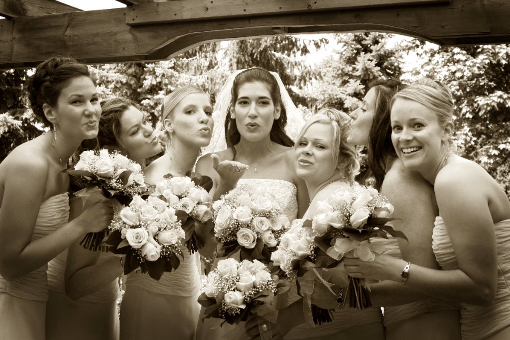 Bride and bridesmaids blowing kisses under wooden pergola, sepia toned