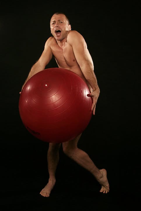 Shirtless male model holding red exercise ball, dramatic side-lit studio portrait