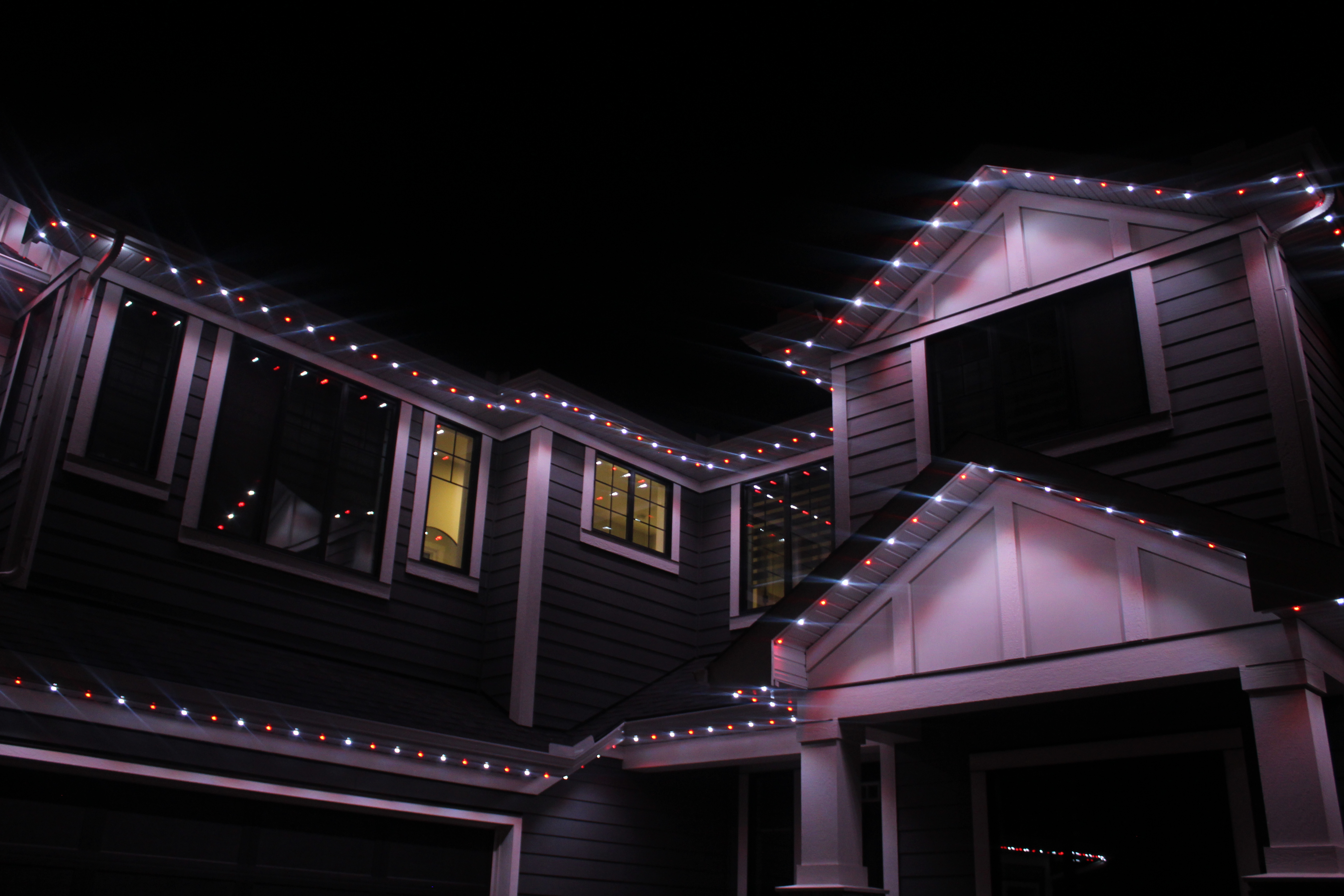 Red and white permanent LED lights outlining roof edges and gables on a dark siding home