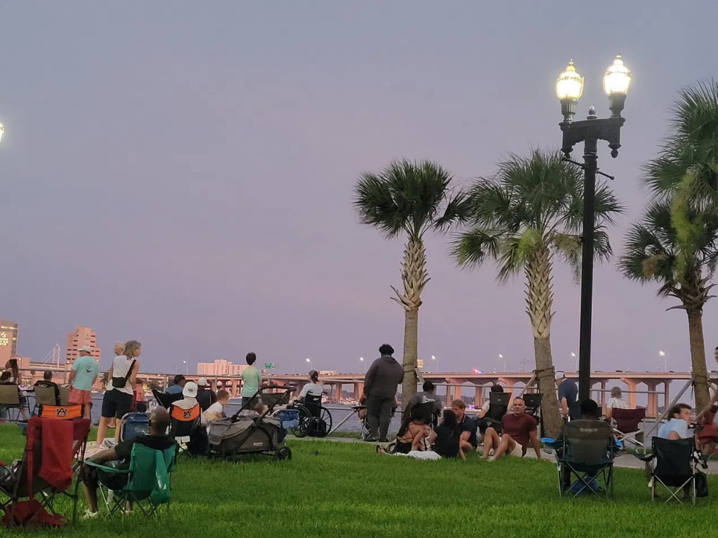 People gathered at a waterfront park at dusk overlooking a lit bridge