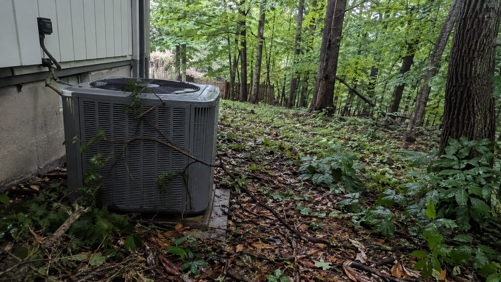 Gray HVAC condenser unit on a concrete pad next to a siding wall on a wooded lot with fallen leaves — Newington VA HVAC service