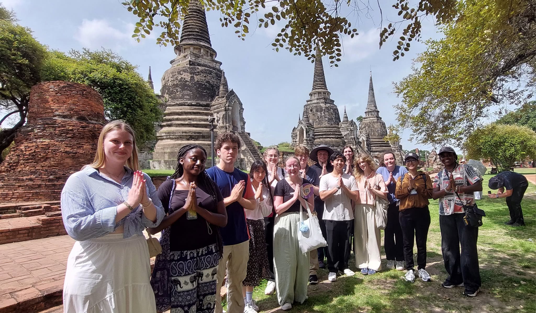 Group of fifteen people posing in wai gesture before ancient brick stupas at Ayutthaya