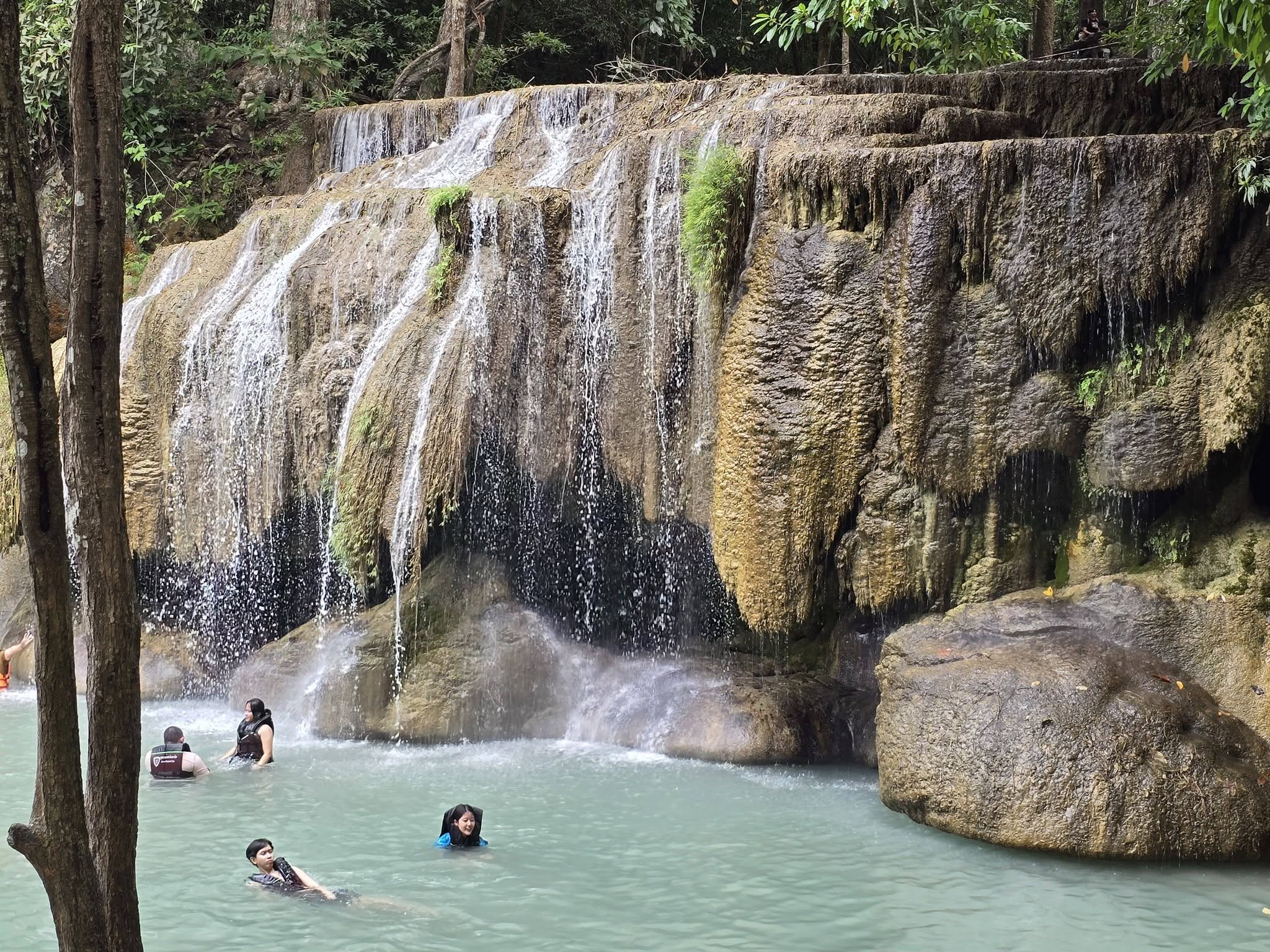 Multi-tiered jungle waterfall with people swimming in the turquoise basin below
