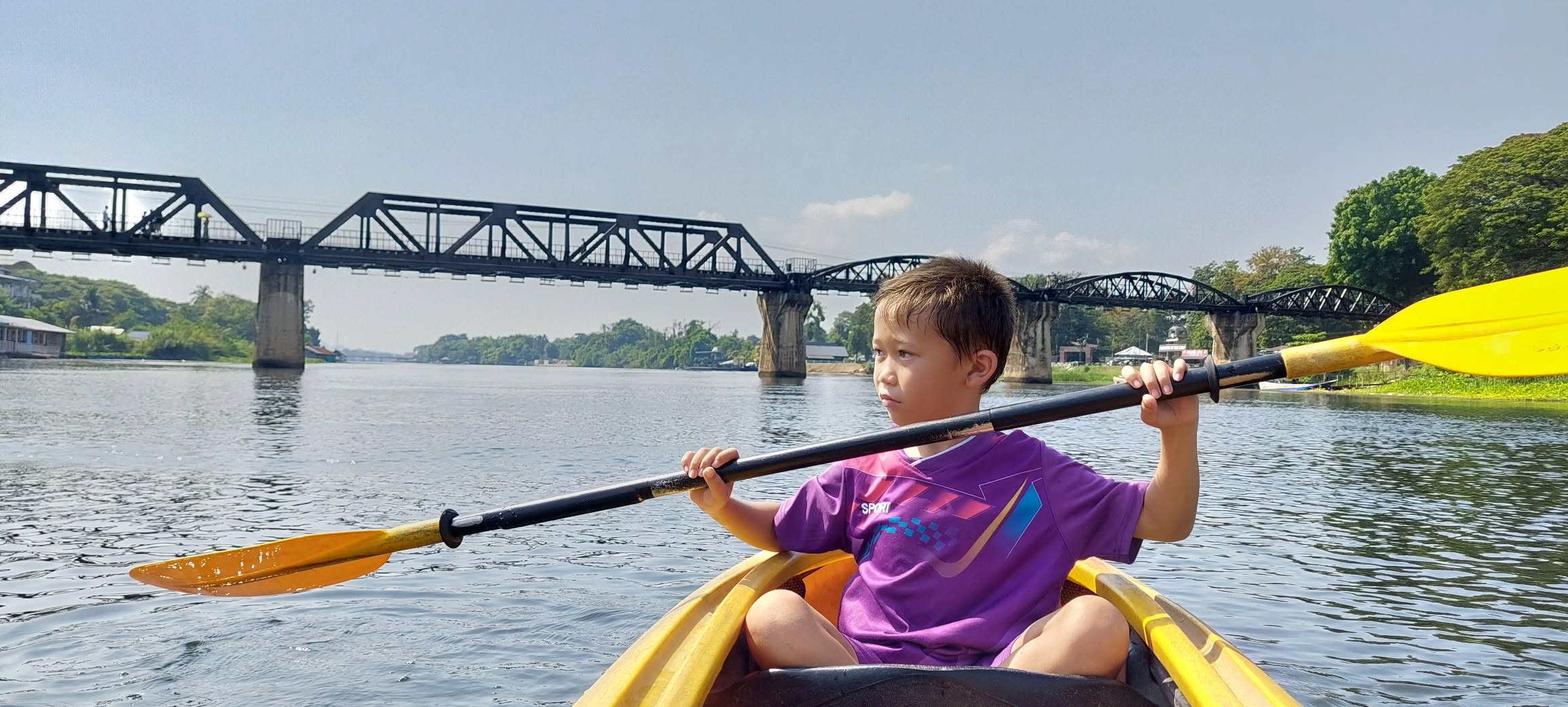 Young boy in a yellow kayak on the river with the historic Bridge over the River Kwai in background