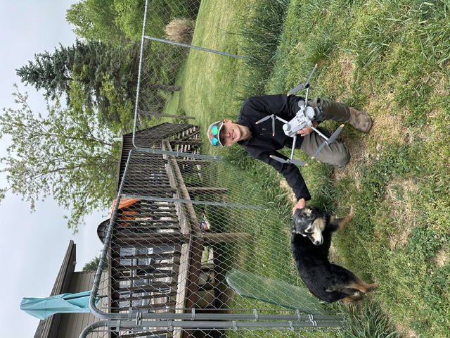 Drone operator kneeling with a dog and drone in hand outdoors
