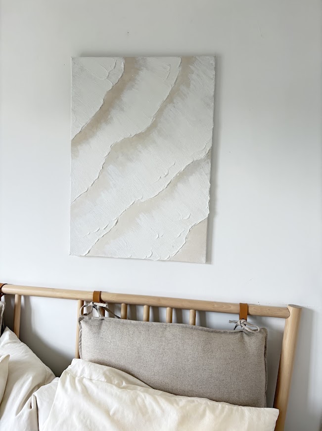 Textured white and beige abstract painting hanging above a wooden bed with linen bedding in a bright minimalist bedroom