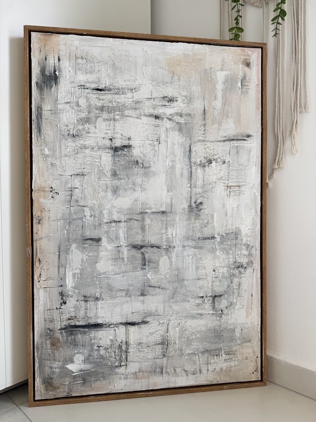 Textured abstract painting in white, grey, black and warm beige tones with heavy impasto brushwork in a wooden frame, leaning against a wall.