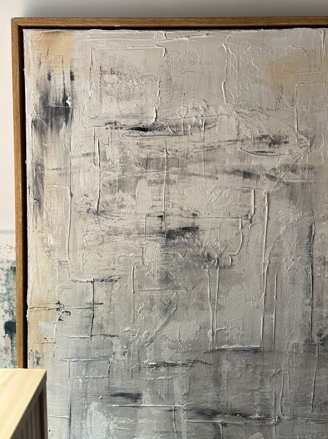 Textured abstract painting in white, grey, black and warm beige tones with heavy impasto brushwork in a wooden frame, leaning against a wall.