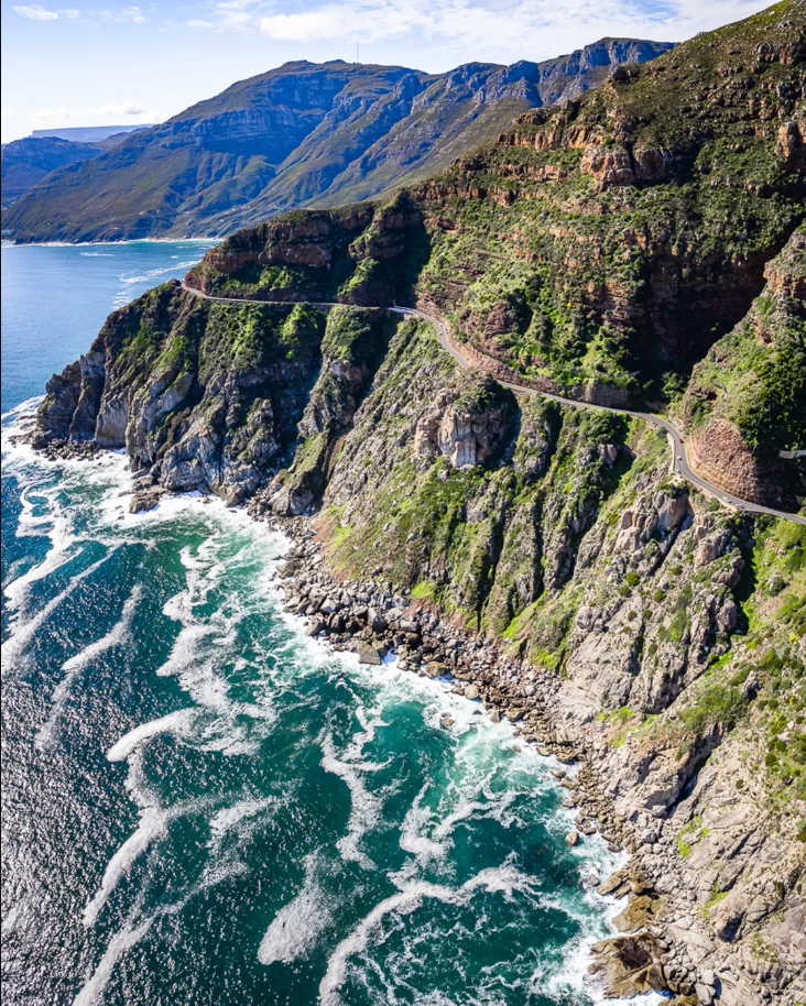 A breathtaking aerial view of Chapman's Peak Drive on the Cape Peninsula, where the winding coastal road hugs steep cliffs that plunge into the turquoise Atlantic Ocean. The rugged mountain terrain, lush greenery, and swirling sea foam create a dramatic contrast, showcasing one of South Africa's most iconic engineering marvels and sightseeing routes.