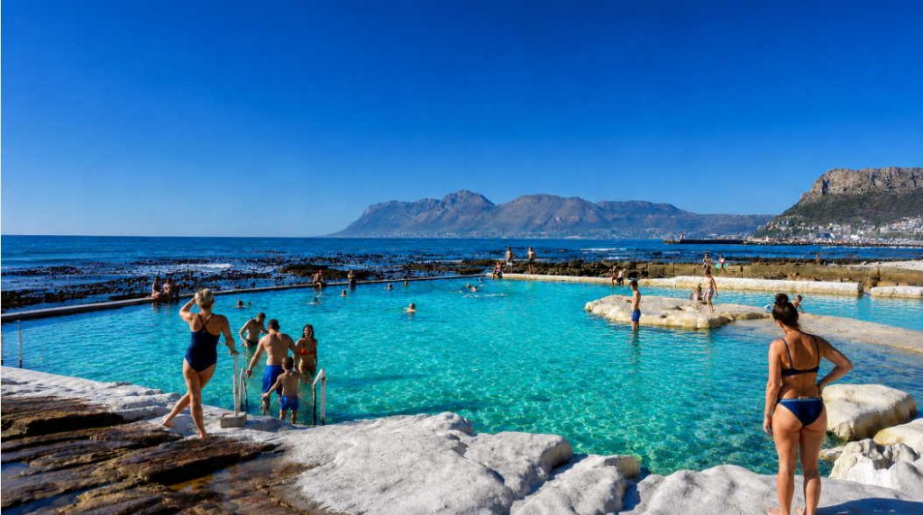 Dalebrook Tidal Pool in Kalk Bay, Cape Town, with crystal-clear turquoise water, swimmers, False Bay coastline and dramatic mountain scenery on a sunny day.