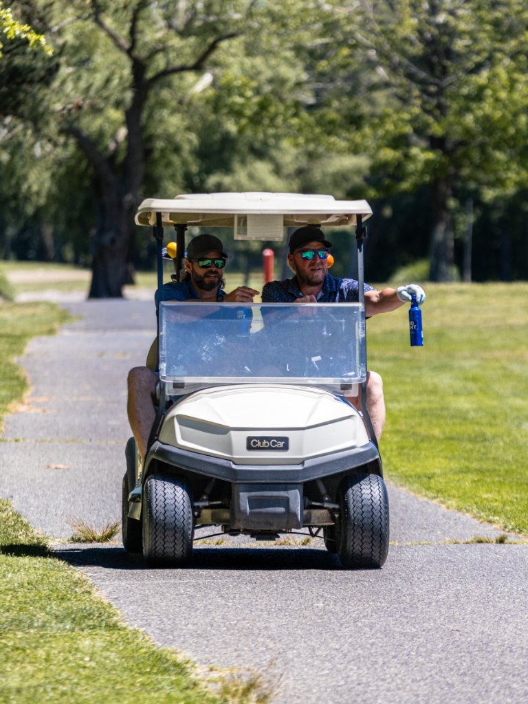Two men in a beige golf cart, one holding up a Bud Light