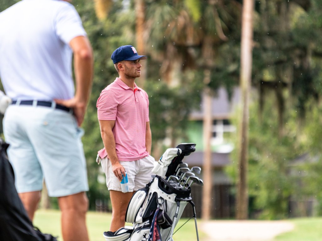 Young golfer in red polo standing beside his bag watching the action