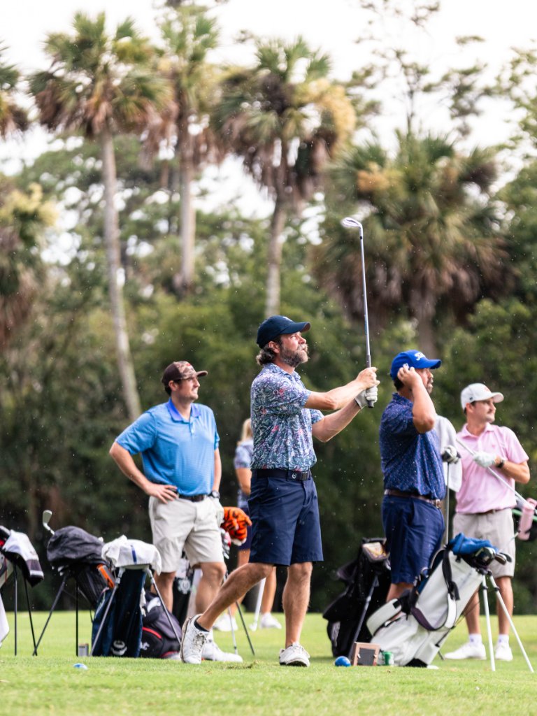 Golfer following through on an iron shot with palm trees and group watching behind