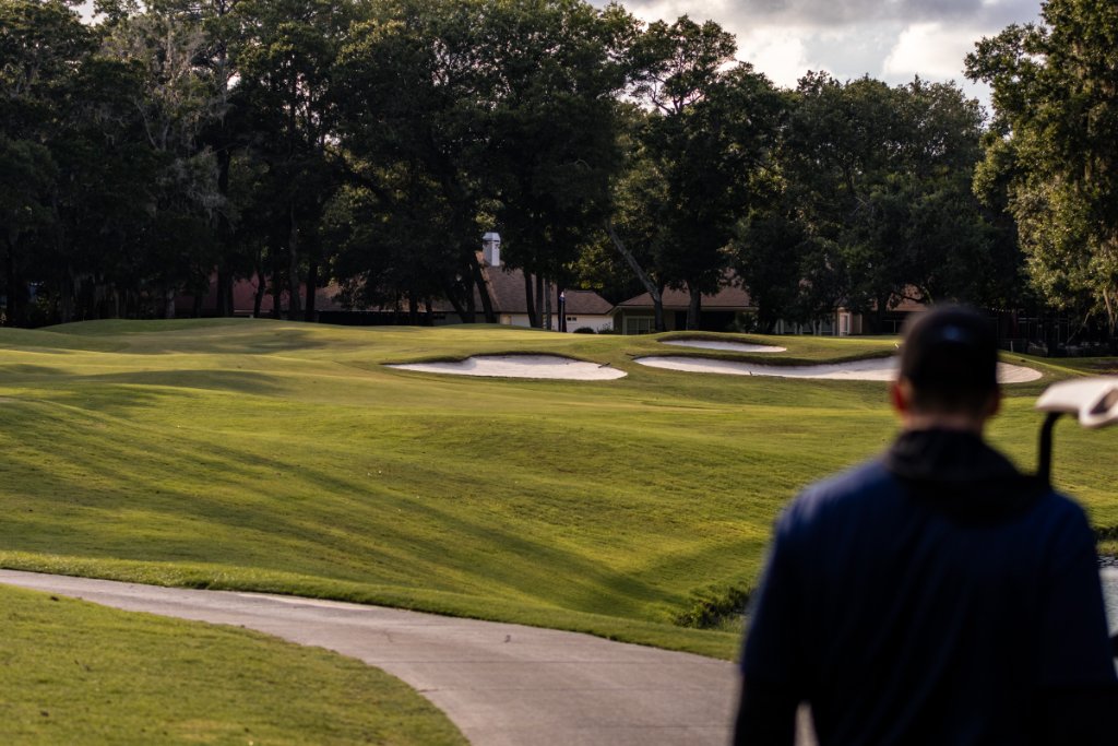 Golfer standing at cart looking out over fairway with bunkers and clubhouse in background
