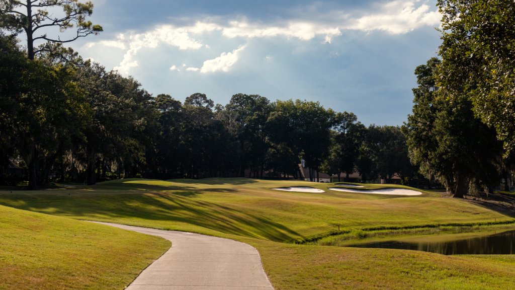 Cart path curving toward a green with bunkers and towering oaks under blue sky