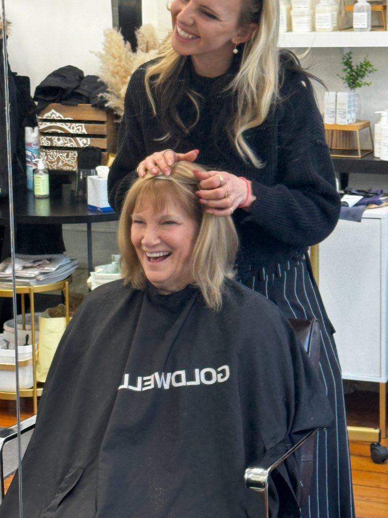Stylist styling a client's blonde hair, both smiling at the salon