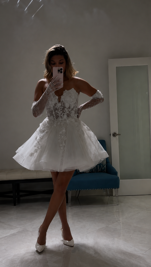 Bride trying on short lace floral applique wedding dress with off-shoulder sleeves and tulle skirt during bridal fitting at Boynton Beach boutique, wedding gown alterations