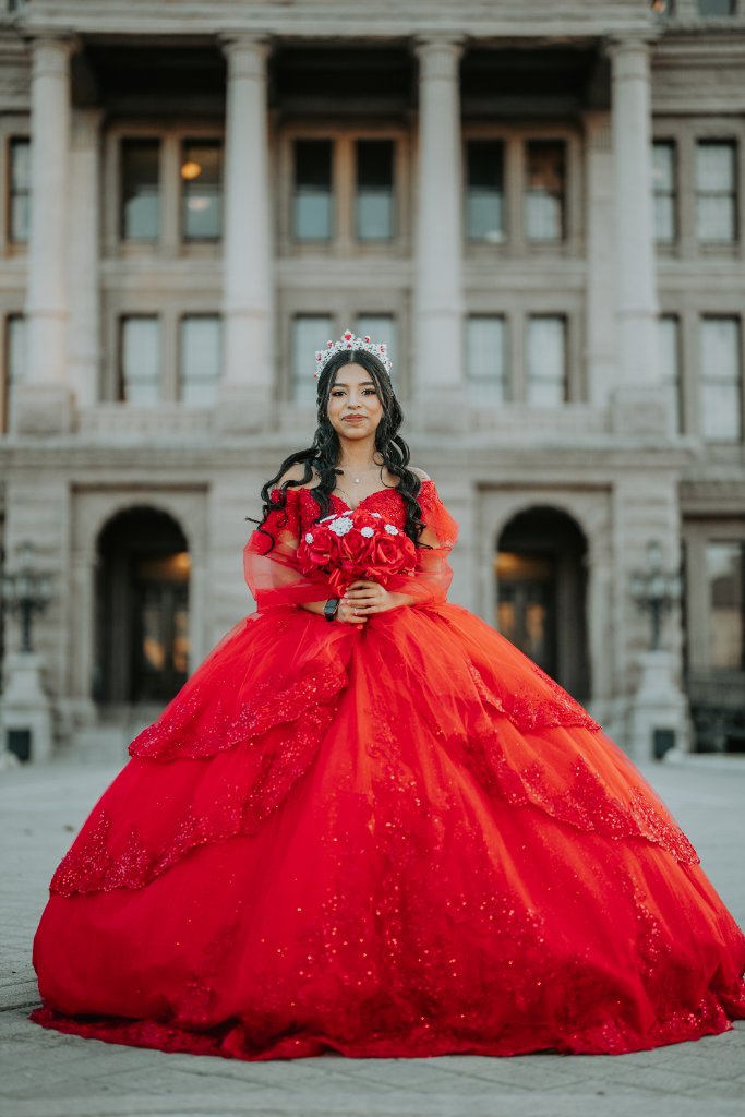 Quinceañera in bright red gown holding roses before stone columns