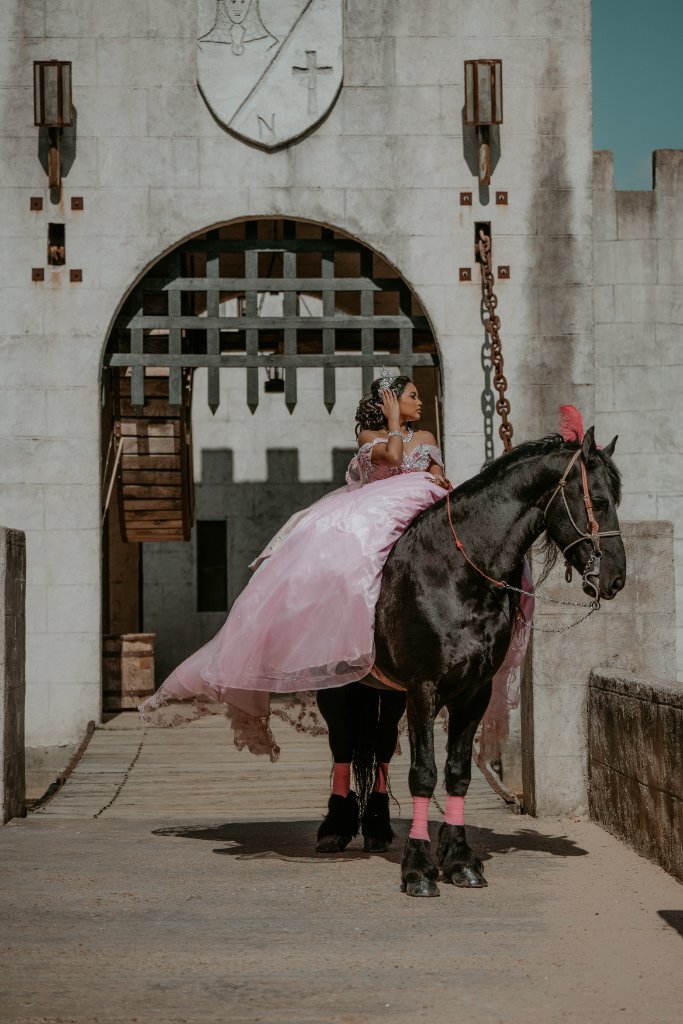 Quinceañera in pink gown seated on a black horse before a castle gate