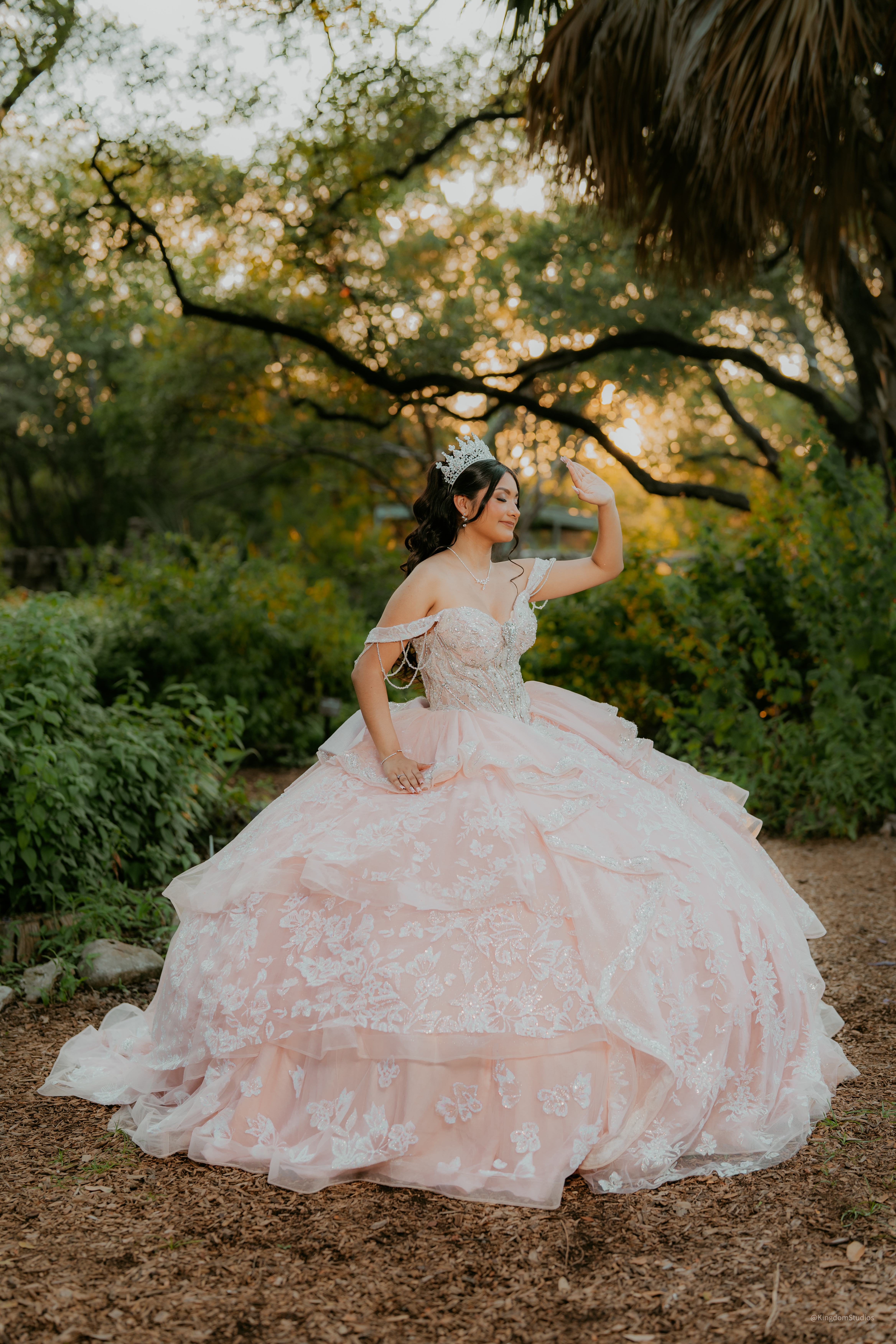 Quinceañera in pale pink gown shielding her eyes in golden forest light