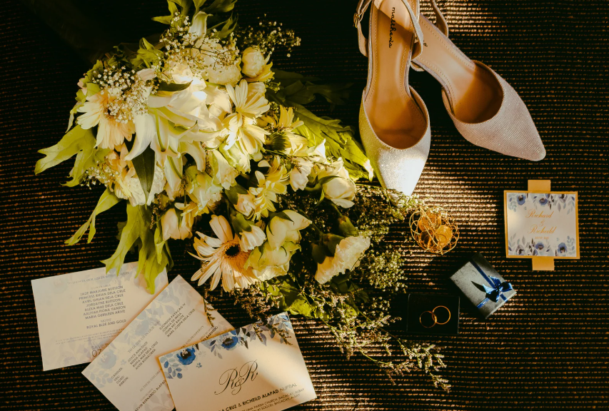 Flat lay of bridal stationery, flowers, shoes, and wedding bands on a dark surface