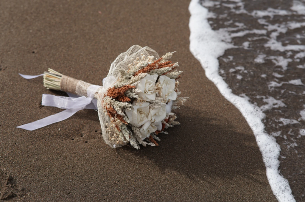Bridal bouquet with cream roses and dried grasses resting on wet beach sand
