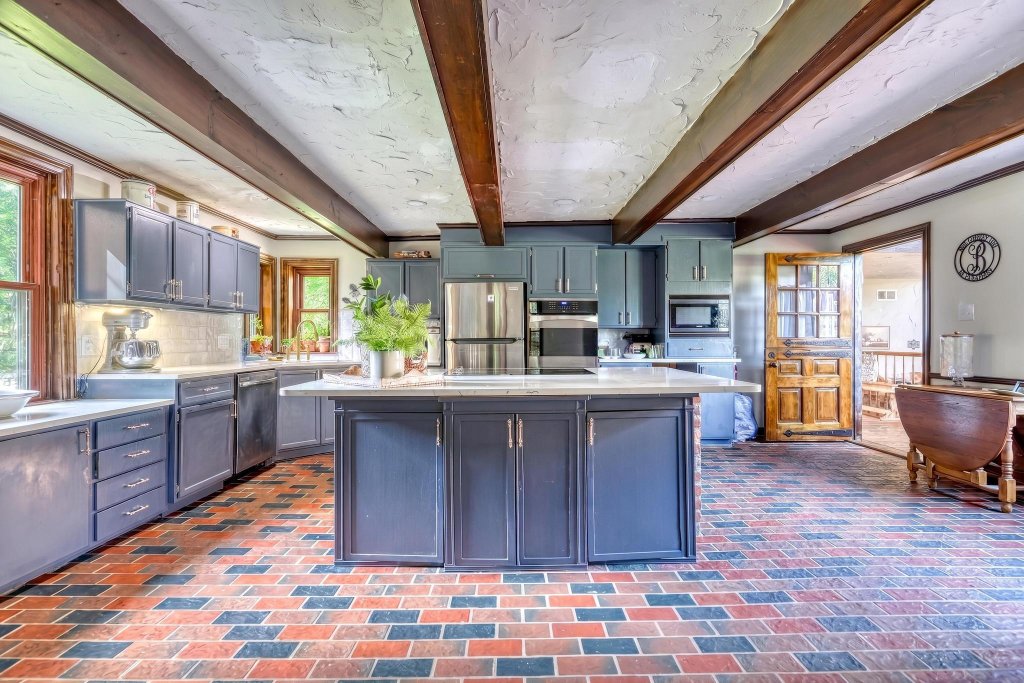 Rustic chef's kitchen with blue-grey cabinetry, brick-tile floor, and exposed beams